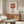 May include: Framed Andy Warhol-style Campbell's tomato soup can print hanging above a mid-century modern walnut credenza in a minimalist living room. The vintage pop art poster features a red and white soup can design in a thin brown frame. Room styled with cream upholstered chair, brass floor lamp, oval glass coffee table, and neutral beige walls with natural lighting.