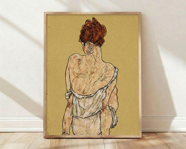 May include: Framed art print showing a figure from behind with reddish-brown hair in an updo, wearing a white undergarment against a golden yellow background. The drawing style features expressive line work and muted earth tones. The wooden frame sits on light wooden flooring against a white wall with soft natural lighting casting shadows.