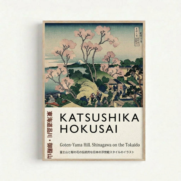 A framed Japanese woodblock print showing cherry blossom trees in pink bloom overlooking a scenic landscape with Mount Fuji in the distance. The artwork features traditional ukiyo-e style with soft green and blue tones depicting rolling hills, small buildings, and people gathered below the flowering trees. Text at the bottom reads "Katsushika Hokusai" and "Goten-Yama Hill, Shinagawa on the Tokaido" with Japanese characters. The print is displayed in a light wooden frame against a white w