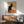 May include: Vintage French poster featuring black cat with yellow eyes sitting upright, ornate decorative background halo, red and yellow text reading "Tournée du Chat Noir de Rodolphe Salis." Art nouveau style advertisement displayed in brown frame on white wall above wooden sideboard. Mid-century modern room setting with sage green upholstered armchair, small table lamp, and coffee table visible.