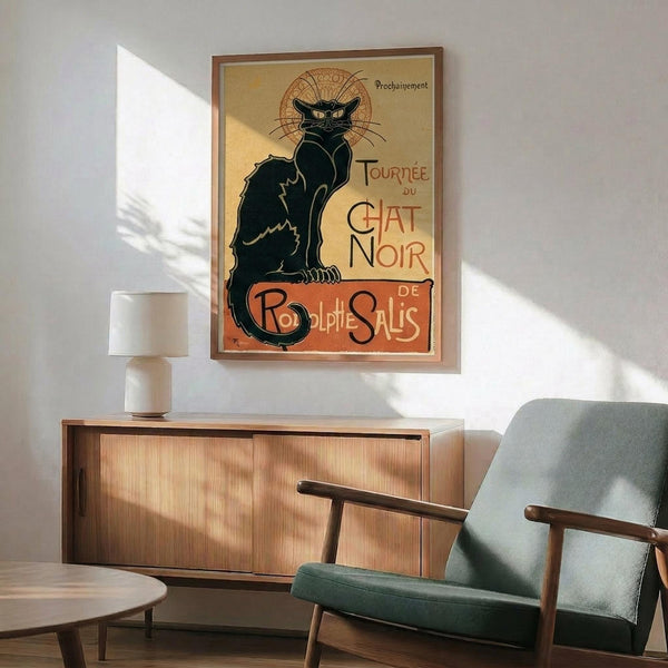 May include: Vintage French poster featuring black cat with yellow eyes sitting upright, ornate decorative background halo, red and yellow text reading "Tournée du Chat Noir de Rodolphe Salis." Art nouveau style advertisement displayed in brown frame on white wall above wooden sideboard. Mid-century modern room setting with sage green upholstered armchair, small table lamp, and coffee table visible.