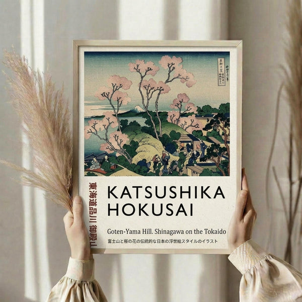 May include: Person holding framed Japanese woodblock print featuring pink cherry blossom trees, Mount Fuji in background, and people walking along hillside path. Print shows traditional ukiyo-e art style with soft blues, greens, and pink tones. Light wooden frame against neutral curtained background with dried pampas grass visible on left side.