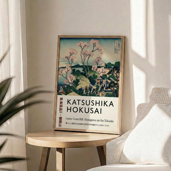 May include: Framed Japanese woodblock print poster featuring cherry blossoms, rolling hills, and traditional buildings in soft pink, green and blue tones, displayed on a light wood round side table next to a beige upholstered chair with decorative pillow, plant leaves visible in corner, natural sunlight casting shadows on white wall