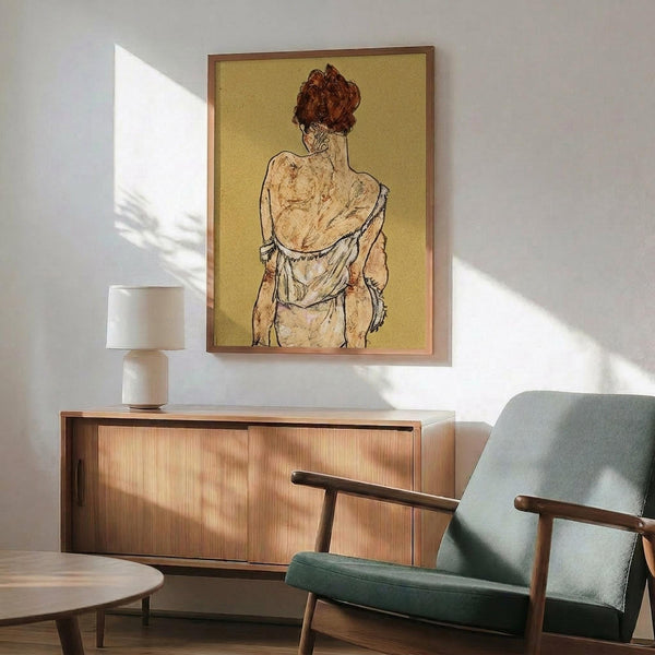 May include: Framed art print of a figure with reddish hair viewed from behind against yellow background, displayed above wooden sideboard with table lamp in modern living room setting with mint green upholstered chair and natural lighting casting shadows on white wall.