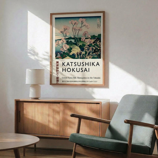 May include: Japanese woodblock print poster in wooden frame showing cherry blossoms and mountain landscape in pink, blue and green tones. Displayed above mid-century modern wooden sideboard with white ceramic lamp. Mint green upholstered armchair with wooden frame sits nearby. Warm sunlight creates geometric shadows on white wall in minimalist room setting.