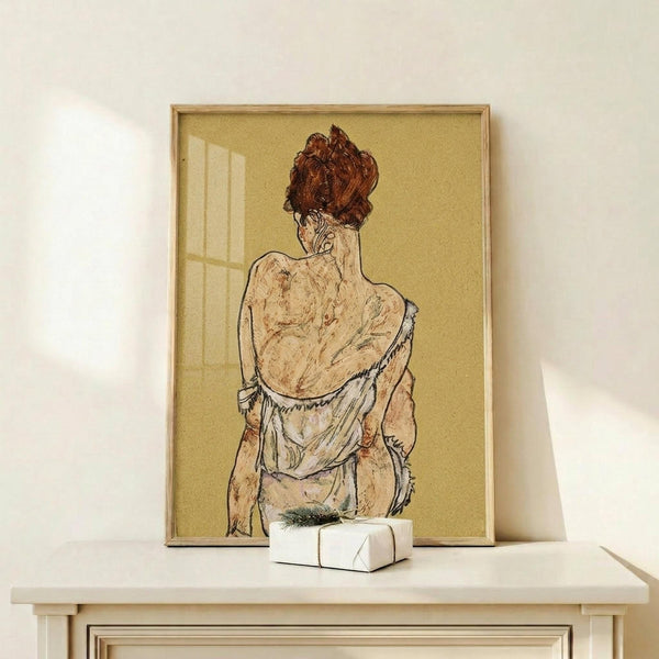 May include: Framed reproduction of an expressionist drawing showing a seated figure from behind with reddish-brown hair, wearing draped white fabric against a golden yellow background. The artwork is displayed in a light wood frame on a white shelf next to a small wrapped gift box, creating a minimalist home decor scene with soft natural lighting.