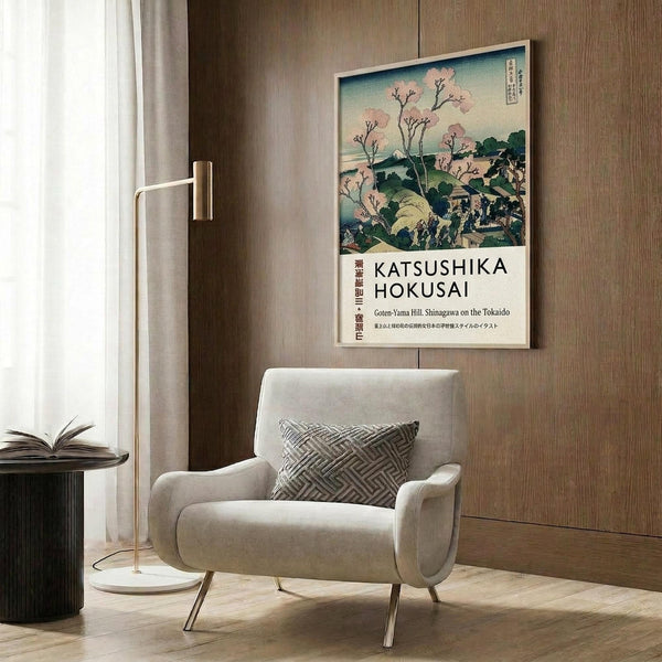 May include: Beige armchair with geometric pillow beneath framed Japanese woodblock print showing cherry blossoms and Mount Fuji in pink, blue and green tones. Modern brass floor lamp and dark round side table with open book nearby. Wood-paneled wall background with natural lighting from window curtains.