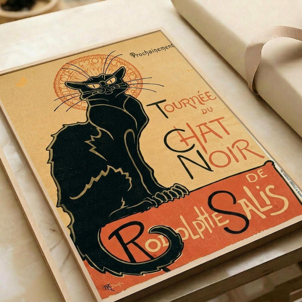 May include: Vintage French poster reproduction featuring a black cat silhouette with yellow eyes against warm beige and orange background, displayed in white matted frame on white surface. The Art Nouveau style advertisement shows "Tournée du Chat Noir de Rodolphe Salis" text in red lettering with decorative circular design behind the cat's head.