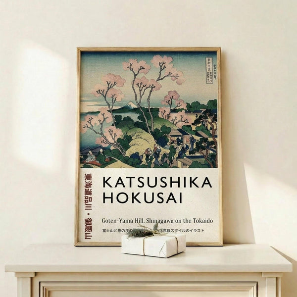 May include: A framed reproduction of a Japanese woodblock print featuring cherry blossom trees in pink bloom overlooking a landscape with Mount Fuji in the distance. The artwork displays traditional ukiyo-e style with soft blues, greens, and pink tones. Below the image is white text reading "KATSUSHIKA HOKUSAI" and "Goten-Yama Hill, Shinagawa on the Tokaido" with Japanese characters on the left side. The wooden-framed poster leans against a white wall on a light wood shelf next to a sma