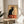May include: Vintage French art nouveau poster featuring a black cat with bright yellow eyes sitting upright against cream background with ornate orange circular design. Red text reads "Tournée du Chat Noir de Rodolphe Salis." Displayed in thin wooden frame on round wooden side table next to beige upholstered chair with white throw pillow. Soft natural lighting creates shadows on neutral wall with plant leaves visible.