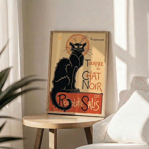 May include: Vintage French art nouveau poster featuring a black cat with bright yellow eyes sitting upright against cream background with ornate orange circular design. Red text reads "Tournée du Chat Noir de Rodolphe Salis." Displayed in thin wooden frame on round wooden side table next to beige upholstered chair with white throw pillow. Soft natural lighting creates shadows on neutral wall with plant leaves visible.