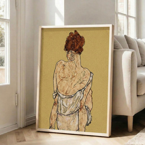 May include: Framed art print showing a figure from behind with reddish-brown hair in an updo, wearing draped clothing rendered in earthy tones against a golden yellow background. The drawing style features expressive line work and muted colors. The print is displayed in a light wooden frame, leaning against a beige couch in a bright room with white walls and large windows.