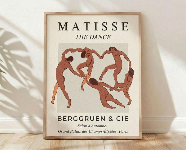 May include: Framed art print featuring Henri Matisse's "The Dance" with five nude figures in terracotta red tones holding hands in a circle against cream background. Black text reads "MATISSE THE DANCE" and "BERGGRUEN & CIE" with Paris gallery details. Light wood frame sits on wooden floor against white textured wall with palm leaf shadows casting across the scene.