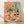 May include: Framed art print leaning against white wall on light wood floor. Artwork shows colorful still life scene with wildflower bouquet in glass vase, wine bottle and glass, assorted foods on plates including bread, cheese, and fruits arranged on peachy pink table. Matisse-inspired style with bold colors, simplified forms, and loose brushwork. Light wood frame, soft natural lighting creates diagonal shadows on textured white wall.