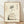 May include: Framed poster print featuring a minimalist ink wash painting of a plump, contented cat sitting with closed eyes and whiskers, rendered in soft black brushstrokes on cream textured paper. Japanese calligraphy characters appear above the cat. The artwork is displayed in a light wood frame, leaning against a white wall on pale wooden flooring with natural window light casting shadows.