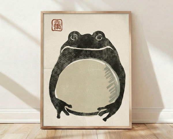 May include: Framed art print leaning against white wall on light wood floor. The poster features a stylized black frog with large round eyes and pale cream belly, rendered in traditional Japanese woodblock print style. A red square seal with characters appears in the upper left corner. The artwork is displayed in a thin natural wood frame against cream-colored paper background.