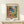 May include: Framed vintage-style poster featuring Mediterranean coastal scene with coral-pink buildings, palm trees, and turquoise water viewed through green shutters. Matisse and Nice text visible at top and bottom. Natural wood frame sits on light wooden surface next to small white wrapped gift with evergreen sprig against cream wall.