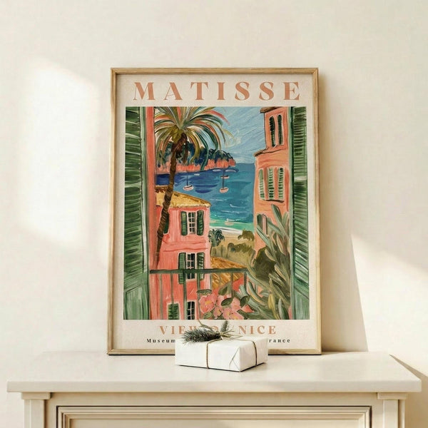May include: Framed vintage-style poster featuring Mediterranean coastal scene with coral-pink buildings, palm trees, and turquoise water viewed through green shutters. Matisse and Nice text visible at top and bottom. Natural wood frame sits on light wooden surface next to small white wrapped gift with evergreen sprig against cream wall.