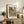 May include: Framed Matisse poster featuring Mediterranean coastal scene with coral-pink buildings, green shutters, palm trees, and blue water view. Print displayed on rustic wooden console alongside fern plant, stacked books, ceramic vase, and table lamp in warm neutral living room setting.