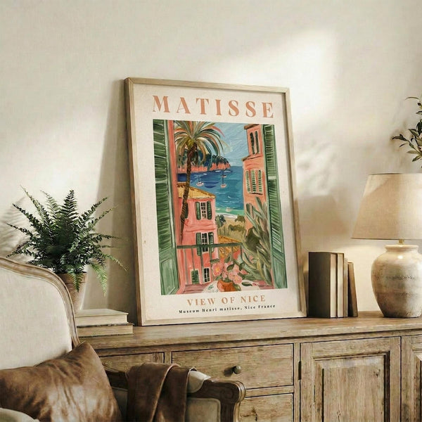 May include: Framed Matisse poster featuring Mediterranean coastal scene with coral-pink buildings, green shutters, palm trees, and blue water view. Print displayed on rustic wooden console alongside fern plant, stacked books, ceramic vase, and table lamp in warm neutral living room setting.