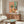 May include: Colorful still life artwork hanging above wooden credenza in modern living room. Painting features vibrant coral background with vase of wildflowers, wine bottle, plates of food, and fruit arranged on pink table. White sculptural chair and glass coffee table visible in foreground. Artwork displayed in thin wooden frame against white wall.