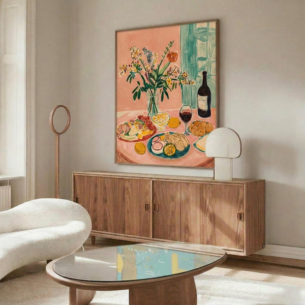 May include: Colorful still life artwork hanging above wooden credenza in modern living room. Painting features vibrant coral background with vase of wildflowers, wine bottle, plates of food, and fruit arranged on pink table. White sculptural chair and glass coffee table visible in foreground. Artwork displayed in thin wooden frame against white wall.