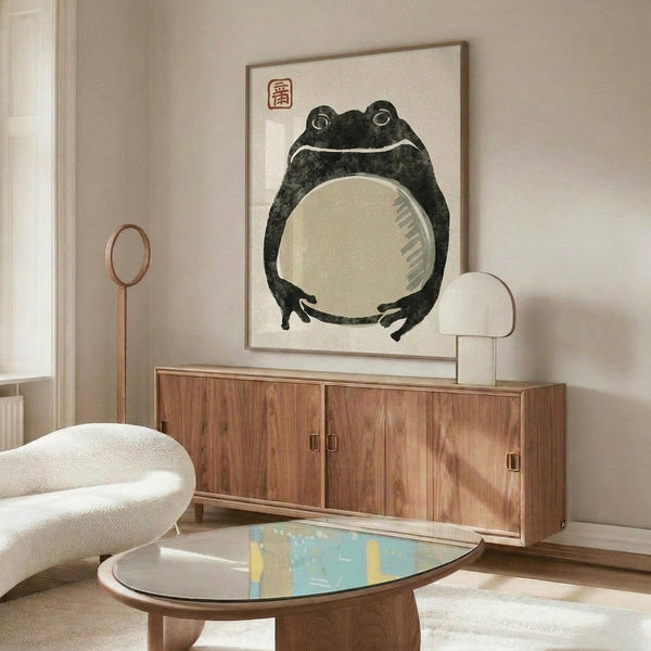 May include: Japanese-style frog artwork print in thin wood frame hanging above mid-century walnut credenza with sliding doors. Dark green frog with large white belly and red square seal stamp on cream background. Modern living room setting with white curved chair, oval glass coffee table, and warm natural lighting creating shadows on neutral walls.