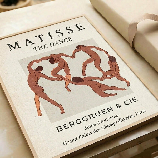 May include: Matisse "The Dance" art print in warm cream tones showing five simplified human figures holding hands in a circle dance. The poster features "MATISSE THE DANCE" at top and "BERGGRUEN & CIE Salon d'Automne Grand Palais des Champs-Élysées, Paris" at bottom. Print sits on neutral fabric surface beside rolled paper, photographed at an angle showing wooden frame edge.