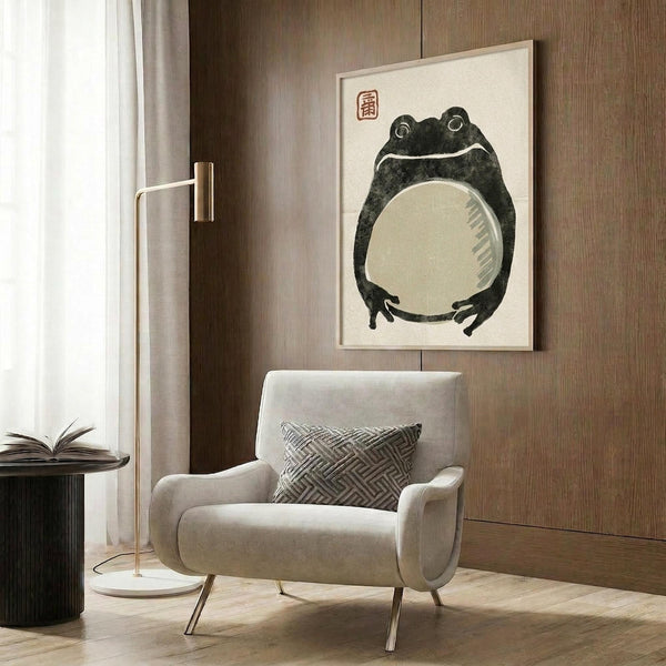 May include: Cream-colored framed poster featuring a minimalist ink wash painting of a dark green frog with large round eyes and pale belly, accompanied by red Japanese characters. Shown mounted on wood-paneled wall above a beige upholstered armchair with geometric patterned throw pillow, alongside a brass floor lamp and side table with open book.