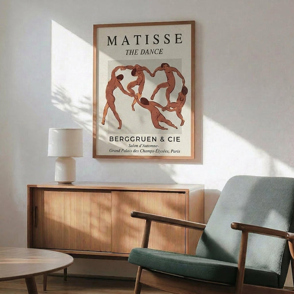 May include: Framed Matisse poster showing five nude figures dancing in a circle, rendered in earthy orange-red tones against cream background. Exhibition text reads "MATISSE THE DANCE" and "BERGGRUEN & CIE" with Paris gallery details. Wooden frame hangs on white wall above mid-century modern credenza with table lamp. Teal upholstered armchair with wooden arms sits nearby, creating warm sunlit interior scene with natural shadows.