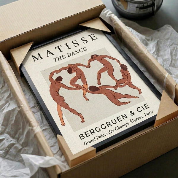 May include: Framed art print of Matisse's "The Dance" showing five red-brown figures dancing in a circle on cream background, displayed in black frame inside cardboard shipping box with white tissue paper and wooden dowels, sitting on white fabric surface next to dark glass object.