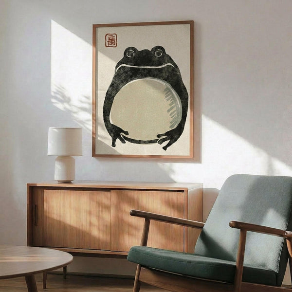 May include: Vintage Japanese frog artwork displayed in wooden frame on white wall above mid-century modern walnut credenza with table lamp, next to sage green upholstered armchair. The print shows a dark charcoal frog with large round belly and small red character seal, rendered in traditional ink wash style. Warm natural lighting creates geometric shadows across the minimalist room setting.