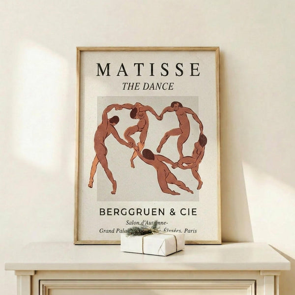 May include: Matisse "The Dance" poster in thin wooden frame on light wood shelf against cream wall. Shows five terracotta-colored dancing figures in a circle on beige background. Black text reads "MATISSE THE DANCE" and "BERGGRUEN & CIE" with Paris gallery details. Small white wrapped gift with evergreen sprig sits beside frame.
