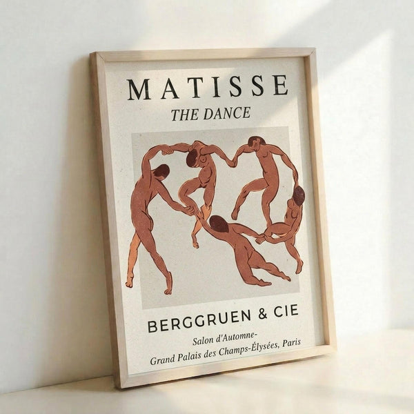 A framed poster featuring five nude figures in reddish-brown tones dancing in a circle against a cream background. The artwork is displayed in a light wood frame and shows text reading "MATISSE THE DANCE" at the top and "BERGGRUEN & CIE" with Paris exhibition details below. The frame leans against a white wall, casting soft shadows.
