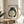 May include: Framed artwork featuring a dark green and black frog with a large pale belly, displayed on a round wooden side table next to a beige upholstered chair. The minimalist print shows traditional Japanese-style brushwork with a red seal stamp in the upper corner. Natural sunlight creates soft shadows across the neutral-toned room setting with a plant visible in the background.