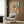 May include: Colorful still life poster print in light wood frame hanging on wood-paneled wall above beige armchair. Artwork features vibrant coral and mint green background with wildflower bouquet in vase, wine bottle, glass, and plates of food on pink tablecloth. Modern living room setting with brass floor lamp, patterned throw pillow, and dark round side table with open book.