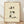 May include: A framed poster print in a light wood frame leaning against a white wall on wooden flooring. The artwork features a cute sitting calico cat with closed happy eyes, pink cheeks, and black and gray patches on cream fur, painted in soft watercolor style. Black Japanese calligraphy characters appear above the cat with a small red seal stamp. The background has a textured cream paper appearance with subtle speckled details.