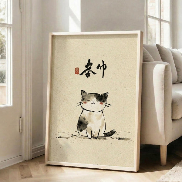 May include: Framed artwork leaning against white wall next to beige sofa, showing watercolor painting of sitting calico cat with black and tan patches on cream textured background, topped with black Japanese calligraphy characters and small red seal stamp, displayed in light wooden frame in bright sunlit room with hardwood floors.