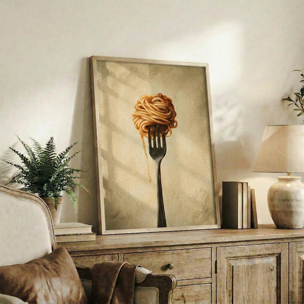 May include: Framed artwork showing pasta twirled on a black fork against a warm, textured beige background, displayed on a rustic wooden sideboard alongside books, a ceramic vase, and a fern plant in a neutral-toned living room setting with natural lighting