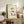 May include: Framed poster with cream background featuring a watercolor-style calico cat with black and white markings sitting peacefully, Japanese calligraphy characters above with red seal stamp. Light wood frame displayed on rustic wooden console table alongside books, ceramic vases, fern plant, and brown leather chair in neutral-toned living room setting.