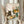 May include: Hands holding a framed art print featuring a gray tabby cat sitting at a checkered tablecloth table with yellow coffee cup, colorful flower bouquet in green vase including sunflowers and pink blooms, and open book. Light wood frame against cream curtains with dried pampas grass visible on left side.