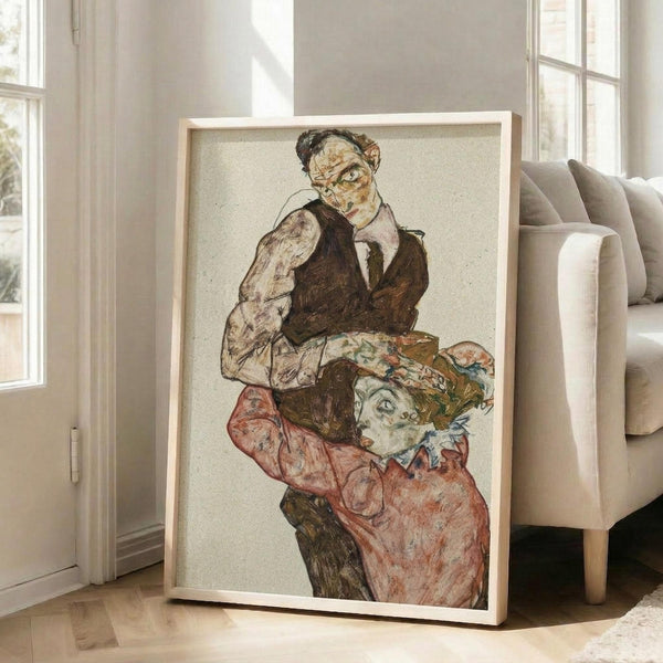 May include: Framed reproduction of an expressionist drawing showing two figures in an intimate embrace, featuring muted earth tones with pops of coral and green. The artwork displays characteristic angular lines and emotional intensity, housed in a light wood frame leaning against white walls in a bright modern living room with beige furniture and large windows.