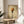May include: Framed poster print showing spaghetti noodles twirled on a black fork against a warm golden beige textured background, displayed on a light wood round side table next to a cream upholstered chair with white pillow, green plant leaves visible, natural window lighting creating soft shadows on white wall
