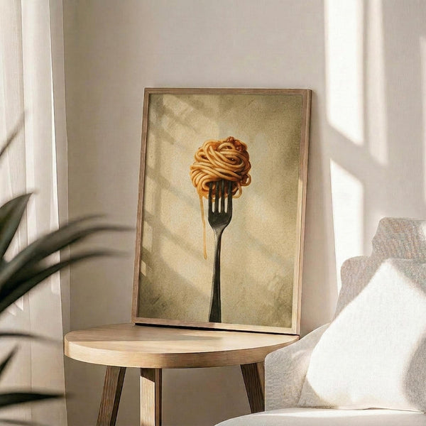 May include: Framed poster print showing spaghetti noodles twirled on a black fork against a warm golden beige textured background, displayed on a light wood round side table next to a cream upholstered chair with white pillow, green plant leaves visible, natural window lighting creating soft shadows on white wall