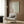May include: Cream-colored framed poster with flowing wave patterns and red Chinese characters mounted on wood-paneled wall above beige upholstered armchair with geometric patterned pillow. Modern brass floor lamp and dark round side table with open book complete the neutral-toned reading nook setting.