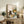 May include: Framed poster print displayed on rustic wooden sideboard in cozy living room setting. Artwork shows black cat sitting at checkered tablecloth table with coffee cup, yellow sunflowers and colorful wildflowers in teal vase. Scene includes wooden chair, open book, warm cottage-style illustration. Room styled with fern plant, cream lamp, books, ceramic vase, beige armchair with brown throw pillow.
