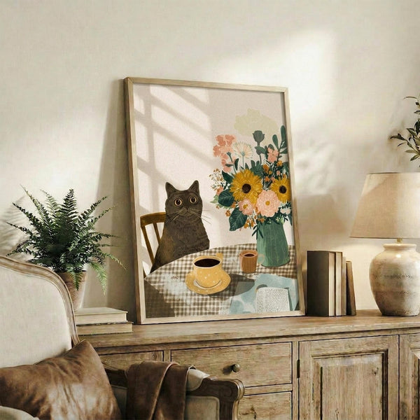 May include: Framed poster print displayed on rustic wooden sideboard in cozy living room setting. Artwork shows black cat sitting at checkered tablecloth table with coffee cup, yellow sunflowers and colorful wildflowers in teal vase. Scene includes wooden chair, open book, warm cottage-style illustration. Room styled with fern plant, cream lamp, books, ceramic vase, beige armchair with brown throw pillow.