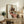 May include: Framed art print displaying an expressionist painting of two figures - a man in dark vest and woman in coral-red dress with floral headpiece, rendered in muted earth tones with bold outlines. Print sits on rustic wooden sideboard alongside fern plant, books, and ceramic vessels in warm-toned living room setting with beige armchair visible.