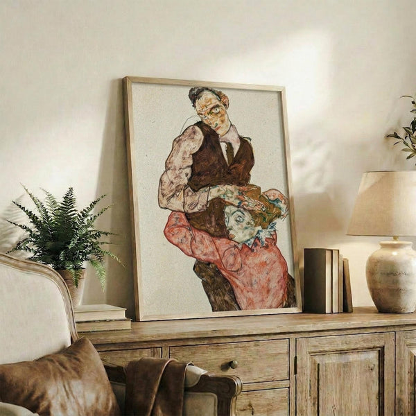 May include: Framed art print displaying an expressionist painting of two figures - a man in dark vest and woman in coral-red dress with floral headpiece, rendered in muted earth tones with bold outlines. Print sits on rustic wooden sideboard alongside fern plant, books, and ceramic vessels in warm-toned living room setting with beige armchair visible.