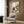 May include: Framed poster print displayed on wood paneled wall above beige upholstered chair with geometric pillow. Artwork shows black cat sitting at checkered tablecloth table with yellow coffee cup, colorful flower bouquet in green vase including sunflowers and pink blooms. Modern reading nook with brass floor lamp, side table with open book, and natural lighting from window with curtains.